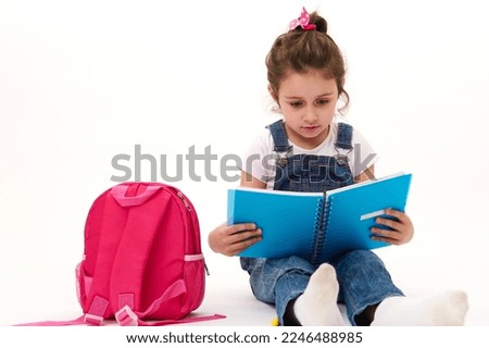 Caucasian lovely child, little girl in blue denim overalls and t-shirt, first grade student reading textbook, sitting near her pink backpack on white background. Copy ad space. Back to school concept Royalty-Free Stock Photo #2246488985