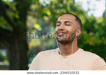 Close-up portrait of sportsman in park, hispanic man jogging in park with eyes closed breathing fresh air and resting, jogging with headphones listening to music and online radio and podcasts. Royalty-Free Stock Photo #2246510533