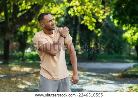 A man injured his shoulder during a fitness class, an African-American man injured himself while jogging in the park, stretches his arm and massages his sore muscles. Royalty-Free Stock Photo #2246510555