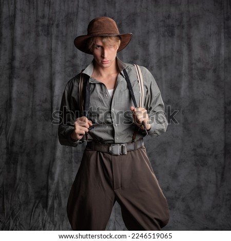 treasure hunter, a hero of a retro-style adventure. A young man in a hat and breeches with suspenders with a whip in his hands. Posing in the studio on a gray background