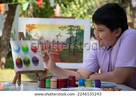 Young Asian boy is looking at example picture on figure stand and using paintbrush to draw various pictures on table outside the room happily, raising teen and adult helps kids school project concept.