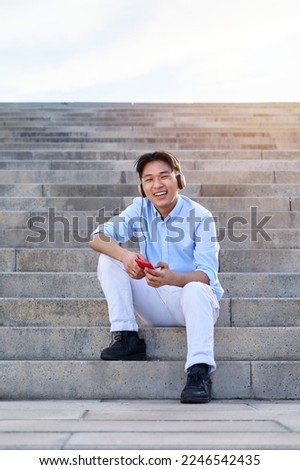 Vertical photo of a cheerful Asian young man wearing headphones looking at camera while sitting on stairs, using mobile app to listen music during break time. Copy spaces image.