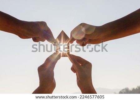 Family Hands holding wooden house toy against blue sunny sky with sun rays. solar power.