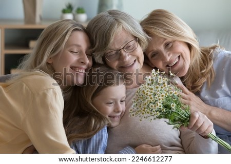 Happy different female generations family portrait. Grandkid woman, mature daughter, pretty cute girl kid hugging cheerful elderly grandma, holding flowers, celebrating mothers day