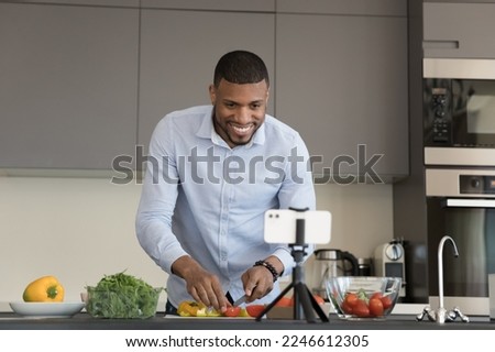 Food blog, on-line recipes. African vlogger record cooking class on smartphone camera, watch tutorial prepare vegetarian dish, cuts fresh paprika and tomato stand in kitchen looks at cellphone screen Royalty-Free Stock Photo #2246612305