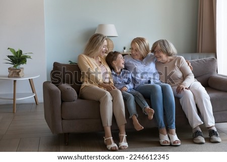 Candid family portrait of happy girls and women of four female generations. Cheerful great grandmother, grandma, mom, daughter kid resting on home sofa together, enjoying meeting, talking, laughing