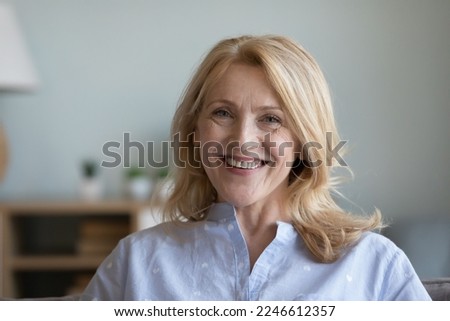 Happy positive senior pretty woman home head shot portrait. Blonde female pensioner sitting on couch, looking at camera with toothy smile, enjoying retirement, promoting elderly beauty, dental care