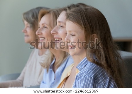 Cheerful cute kid girl standing near young mom, mature granny, old great grandmother, looking away, with tooth gap smile, thinking, laughing. Side portrait with four female generations, family dynasty