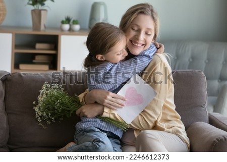Happy peaceful young mom hugging adorable daughter with love, gratitude, holding flowers, greeting card with drawn heart, smiling with closed eyes, laughing, celebrating mothers day