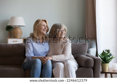 Happy dreamy middle aged daughter woman and old mom sitting close on home sofa, holding hands, looking at window away, discussing future plans, dreaming, thinking, laughing Royalty-Free Stock Photo #2246612379