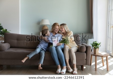 Joyful grandkid girl and adult daughter giving greeting card with heart and flower bunch to happy mature grandmother, sitting home sofa together, hugging, celebrating mothers day