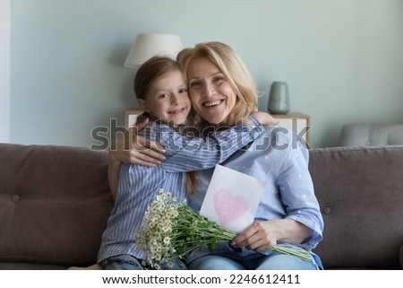 Joyful happy grandma hugging adorable little granddaughter kid girl, holding flowers bouquet, greeting card with heart, looking at camera, smiling, celebrating birthday, mothers day