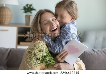 Cheerful cute daughter kid kissing pretty young mom, giving flowers bunch, greeting card due to mothers day, birthday, 8 march, hugging woman with love, tenderness, affection