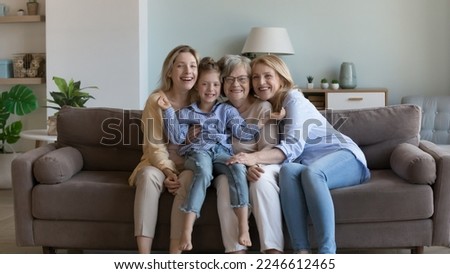 Happy kid girl, young mom, mature grandma, elderly senior great grandmother sitting together on home sofa, holding child in arms on lap, looking at camera, smiling. Four generations family portrait