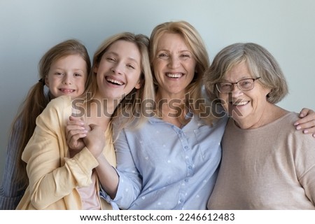 Happy little child, cheerful young mom, middle aged granny, elderly great grandma portrait. Four girls and women of different generations standing at grey wall background, hugging with love, smiling