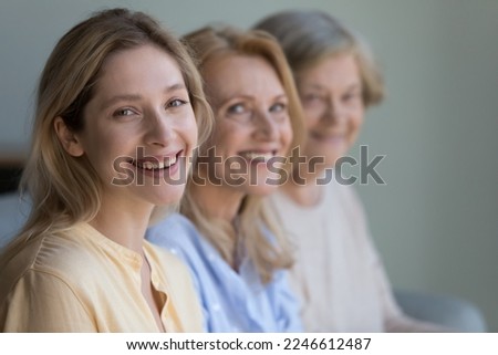 Happy beautiful young adult daughter girl looking at camera, smiling, posing with mature mom and elderly grandma in blurred background. Three female generations family portrait