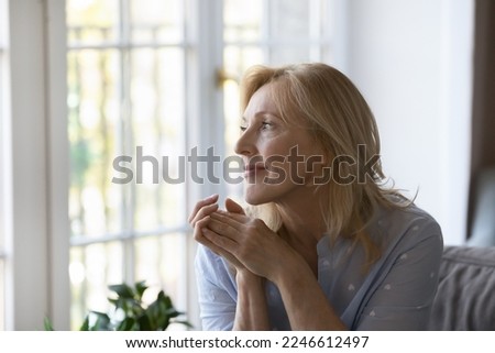 Positive thoughtful pretty middle aged blond woman sitting on home sofa, keeping hands at chin, looking at window away with good thoughts, thinking over future retirement, planning, dreaming