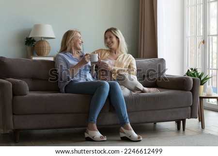 Happy middle aged mom and pretty adult daughter chatting over cup of coffee, enjoying meeting, family leisure, sitting on home couch together, talking, holding mugs of hot drinks. Full length shot Royalty-Free Stock Photo #2246612499