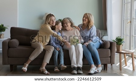 Happy girls and women of four different generations celebrating mothers day, 8 march together, giving flowers to elderly great grandmother, hugging, smiling, laughing on home couch. Banner shot