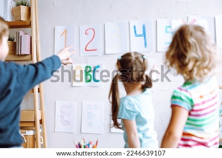 Children stand with back and learn english letters, alphabet and numbers on cards on wall. Kids point at colored numbers on sheets of paper by finger. Reading and counting, study in daycare centre. 