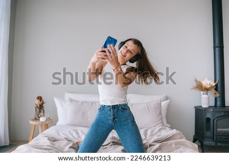 Fit healthy Caucasian young woman in white t-shirt and blue jeans uses headphones makes selfie, home. Pretty hispanic girl makes video call from hotel room enjoys vacations. Youth and travel, vacation