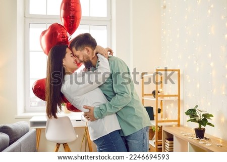 Happy young couple hugging and kissing at home. Husband and wife hugging enjoying tender romantic moment against background of red heart shaped balloons and glowing lights Royalty-Free Stock Photo #2246694593