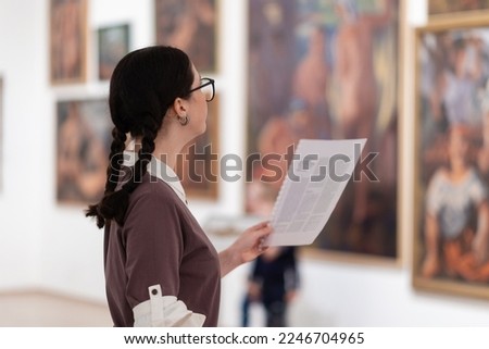 Side view of caucasian pretty young woman wearing glasses holds educational brochure. Defocused pictures in background. Concept of exhibition in gallery.