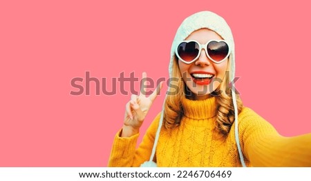 Winter portrait of happy smiling young woman stretching hand for taking selfie with phone wearing yellow knitted sweater, white hat, heart shaped sunglasses on pink background