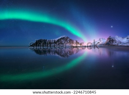 Aurora borealis and beach in Lofoten islands, Norway. Beautiful northern lights. Starry sky with polar lights. Night winter landscape with aurora, sea with sky reflection, city lights, snowy mountains Royalty-Free Stock Photo #2246710845