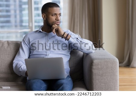 Pensive African guy deep in thoughts, ponders, thinks seated on sofa with laptop staring into distance looks puzzled, uncertain distracted from tech usage, having question or challenge feels undecided Royalty-Free Stock Photo #2246712203