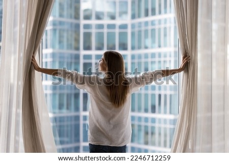 Rear view young woman standing indoors, looking out panoramic window of luxury modern apartment or hotel room opens curtains in morning, enjoying city skyscrapers view, feels happy, welcoming new day Royalty-Free Stock Photo #2246712259