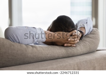 Close up rear view of unknown African single man relaxing leaned on soft cushions of cozy sofa, put hands behind head, enjoy day off at modern skyscraper hotel room, breath fresh air, relish weekend Royalty-Free Stock Photo #2246712331