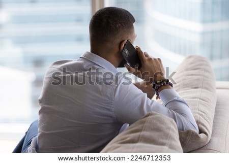 Rear view African man talking on cell phone seated on sofa staring out panoramic window at big city view, communicates to friend or customer services manager, solve business from home. Communication Royalty-Free Stock Photo #2246712353