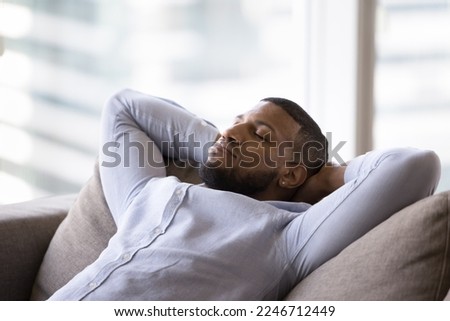 Calm African man rest, take break, daytime nap leaned on cozy soft sofa cushions, closing eyes breathes fresh conditioned air, sleeps inside modern ventilates living room at home or hotel. Relaxation Royalty-Free Stock Photo #2246712449