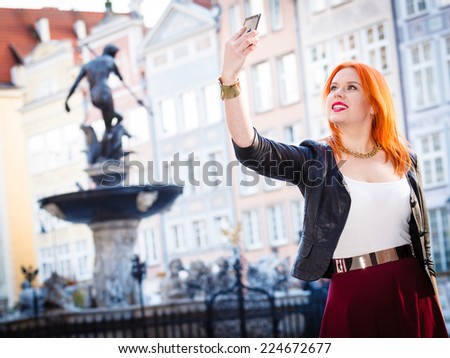 Traveler woman redhair fashion girl taking self picture selfie with smartphone camera outdoors in european city old town Gdansk. Neptune fountain in the background Poland