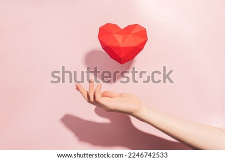 Red volumetric heart made of paper over hand with hard shadow against pink background.