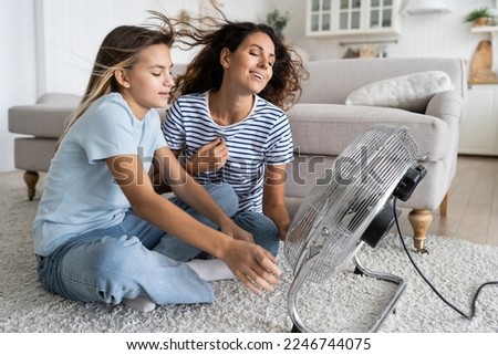 Mother and daughter cooling down near electric fan blowing fresh air, spending time together at home on hot summer days, mom and child sitting on floor in front of ventilator cooler during summer heat Royalty-Free Stock Photo #2246744075