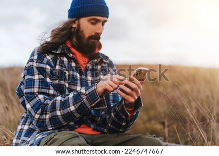Close up of male hand travel photographer in blue jacket checking map on smart phone while sitting against blurred green hills during trekking in autumn countryside on sunset.