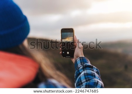 Close up of male hand travel photographer in blue jacket taking pictures on smart phone while sitting against blurred green hills during trekking in autumn countryside on sunset.