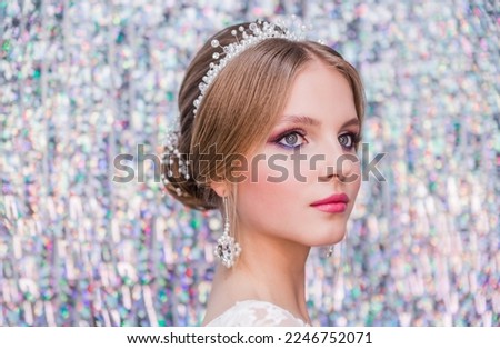 A girl in a white wedding dress with an open back, make-up, a hairstyle in the form of a bunch poses on glitter curtain silver background. Horizontal photo.