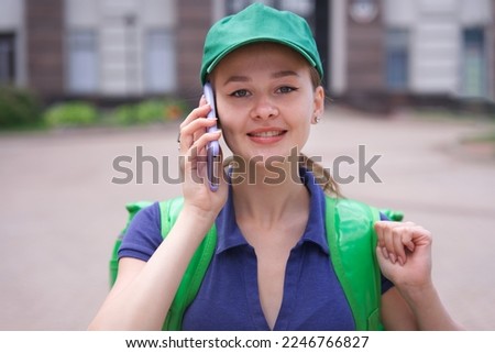 portrait of young beautiful happy courier teenager girl, food delivery woman with green thermo box for food delivering food outdoors in cap and uniform, using cell mobile phone, calling on smartphone