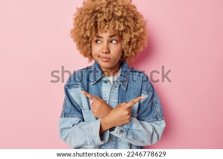 Perplexed curly haired woman points sideways with index fingers shows left and right banner looks questioned by decision dressed in denim jacket isolated over pink background feels hesitant.