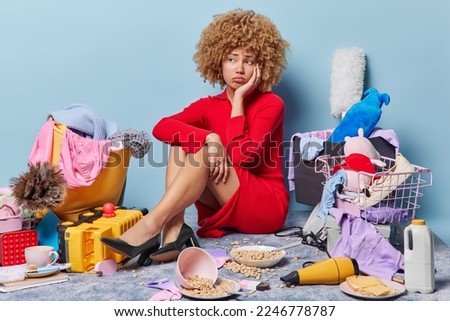Unhappy stressed lady wears red dress and black high heeled shoes sits on floor against different items around upset because of chaos in house looks sadly away isolated over blue background. Royalty-Free Stock Photo #2246778787
