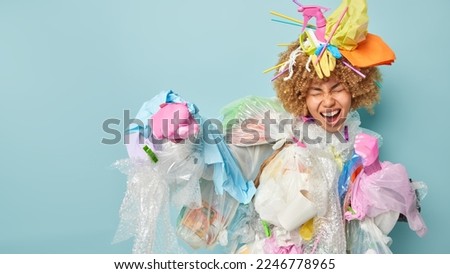 Positive curly haired female volunteer clenches fists pretends fighting against contamination asks not to pollute our planet makes costume from plastic garbage isolated on blue background blank space