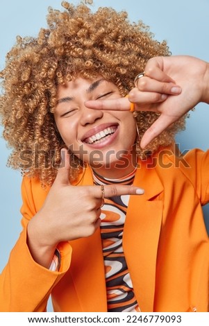 Vertical shot of positive curly haired woman smiles broadly keeps eyes closed makes frame with hands and fingers has happy face dressed in elegant orange jacket isolated over blue background