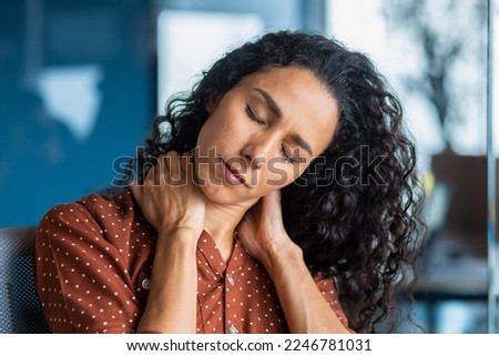 Close-up photo. Tired young hispanic woman. She sits in the office, holds and massages her neck with her hands, closed her eyes, feels pain, tension. Long working overtime day, deadline.