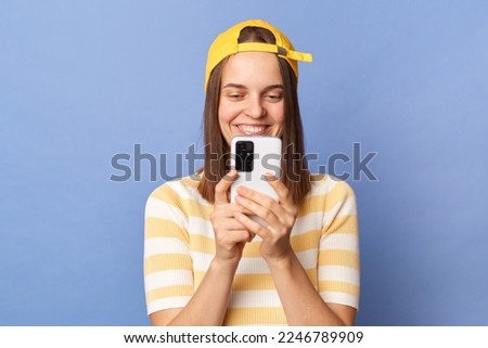 Portrait of smiling teen girl wearing striped T-shirt and baseball cap standing isolated over blue background, holding smartphone, chatting with friends, making photo.