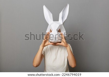 Photo of unknown woman wearing white T-shirt and paper rabbit mask, keeps hands near mouth, screaming loud, making announcement, telling advertisement, standing isolated over gray background.