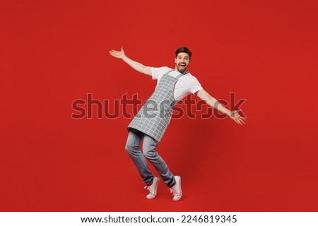 Full body side view happy young housekeeper chef cook baker man wear grey apron look camera with outstretched hands stand on toes lean back isolated on plain red background studio Cooking food concept