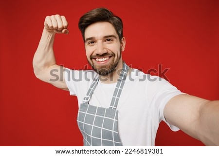 Close up young male housewife housekeeper chef cook baker man wear grey apron doing selfie shot pov on mobile cell phone do winner gesture isolated on plain red color background. Cooking food concept